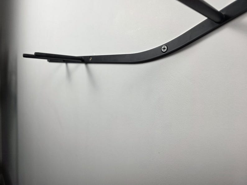 K&ouml;nnte beinhalten: Ein schwarzes Metall-Wandregal mit geschwungenem Design. Das Regal ist an einer wei&szlig;en Wand befestigt und mit Schrauben gesichert. Das Regal hat zwei horizontale Stangen, die sich von der Hauptst&uuml;tze erstrecken.
