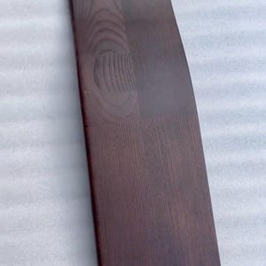 K&ouml;nnte beinhalten: Ein langes, gebogenes Holzbrett mit dunkelbraunem Finish. Die Holzmaserung ist sichtbar, und das Brett scheint aus mehreren zusammengef&uuml;gten Teilen zu bestehen. Das Brett hat eine glatte, polierte Oberfl&auml;che.