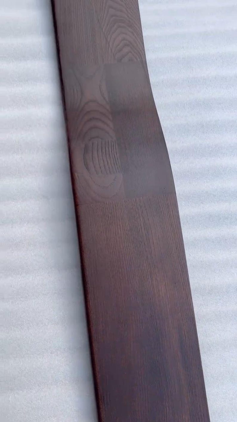 K&ouml;nnte beinhalten: Ein langes, gebogenes Holzbrett mit dunkelbraunem Finish. Die Holzmaserung ist sichtbar, und das Brett scheint aus mehreren zusammengef&uuml;gten Teilen zu bestehen. Das Brett hat eine glatte, polierte Oberfl&auml;che.