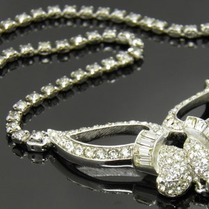 May include: A silver necklace with a large, ornate pendant set with clear rhinestones. The pendant features a delicate, intricate design with multiple loops and curves.