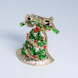 May include: A gold-tone Christmas bell brooch with a green holly and red berry design. The bell has a bow on top and a pin on the back.