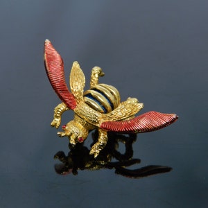 May include: A gold-toned bee brooch with red and gold wings, a striped abdomen, and red gemstone eyes. The brooch is detailed with intricate textures and a polished finish, suitable for adding a touch of nature-inspired elegance to any outfit.