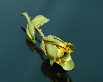 Könnte beinhalten: Eine goldfarbene Rosenbrosche mit strukturierter Oberfläche. Die Rose ist mit einem einzelnen Stiel und Blättern dargestellt.