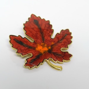 May include: A gold-toned brooch in the shape of a red maple leaf with black veins and an orange centre.