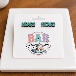 Könnte beinhalten: Ein Paar ohrstecker in Blaugrün mit dem Wort HEARD in Blockschrift, präsentiert auf einer weißen rechteckigen Karte. Die Karte zeigt das Logo BAR Handmade mit einer Berggrafik sowie den Text CUSTOM MADE, PERSONALIZED, CRAFTED.