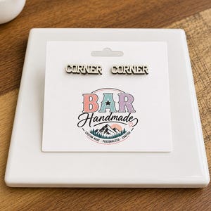 Könnte beinhalten: Zwei silberfarbene Metallanstecknadeln mit dem Wort CORNER in einer fetten Blockschrift. Die Anstecknadeln sind auf einer weißen Anzeigekarte mit dem Text BAR Handmade, Custom Made, Personalized, Crafted und einer Berggrafik befestigt.