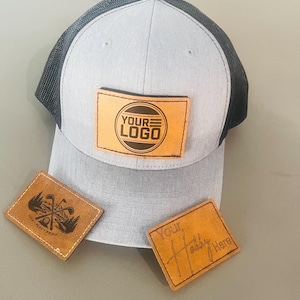 May include: A light grey and black trucker hat with a rectangular tan leather patch featuring the text YOUR LOGO. Two additional tan leather patches are displayed, one with a golf graphic and the text LIVE PLAY ENJOY, and one with the text Your Hobby Here.