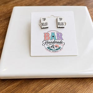 Könnte beinhalten: Ein Paar silberfarbene Ohrstecker in Form kleiner rechteckiger Quittungen, jeweils mit dem schwarzen Aufdruck TIP und $0.00. Die Ohrringe sind auf einer weißen Karte mit dem Logo BAR Handmade und einer Berg-Grafik befestigt.