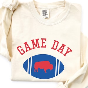 May include: An ivory long-sleeve t-shirt featuring the text GAME DAY in red arched lettering above a blue football graphic containing a red buffalo silhouette. The shirt has a crew neckline and a Comfort Colors brand label at the inner collar.