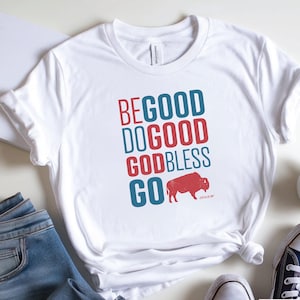May include: White t-shirt with the words "BE GOOD DO GOOD GOD BLESS GO" in red and blue. A red bison graphic is below the text. The shirt is paired with blue jeans and white sneakers.