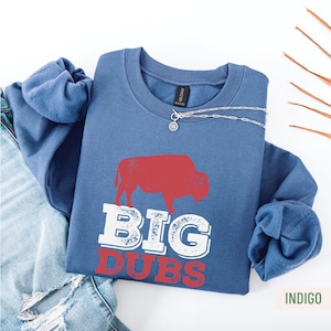 Puede incluir: Sudadera azul índigo con un gráfico de bisonte rojo y las palabras "BIG DUBS" en blanco y rojo. Un collar plateado con un colgante está sobre la sudadera. La sudadera está doblada sobre unos vaqueros azules.