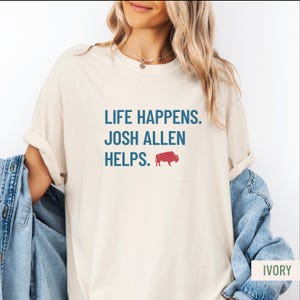 May include: Ivory-colored t-shirt with the text "LIFE HAPPENS. JOSH ALLEN HELPS." in blue, and a red buffalo graphic. The shirt has a relaxed fit and rolled-up sleeves. A denim jacket is partially visible.