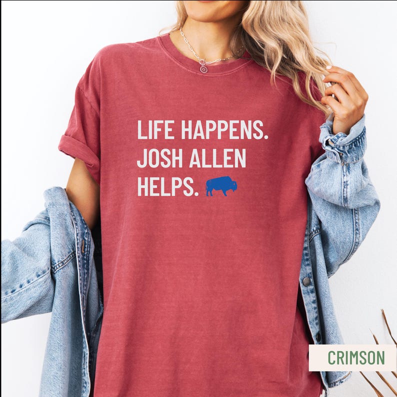 May include: Crimson-colored t-shirt with the text "LIFE HAPPENS. JOSH ALLEN HELPS." in white, and a blue buffalo logo. The shirt has rolled-up sleeves and a casual fit.