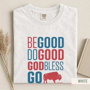 May include: White t-shirt with the words "BE GOOD DO GOOD GOD BLESS GO" in red and blue block letters. A red bison graphic is below the text. The shirt is folded and displayed on a light background.