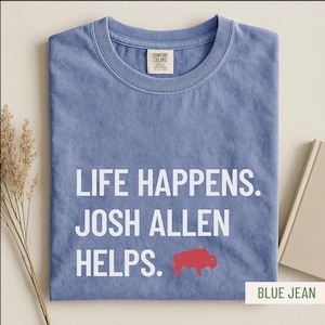 May include: Blue jean colored t-shirt with the text "LIFE HAPPENS. JOSH ALLEN HELPS." in white. A red buffalo graphic is below the text. The shirt has a crew neck and a Comfort Colors tag.
