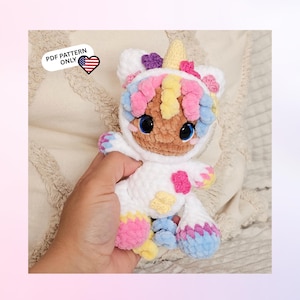 Könnte beinhalten: Ein gehäkeltes Plüschtier in Form eines weißen Einhorn-Kapuzenanzugs mit gelbem Horn, pastellfarbenem rosa, blauem und gelbem lockigem Haar sowie großen blauen Augen. Das Spielzeug hat rosa und gelbe Akzente an Gliedmaßen und Körper. Text: PDF PATTERN ONLY.