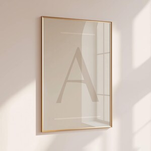 Op de afbeelding: Een ingelijste print met de letter "A" in een zachte beige tint. Het kunstwerk is ingesloten in een dun, goudkleurig frame, wat een minimalistische esthetiek creëert. De print wordt gepresenteerd tegen een neutrale muur, met schaduwen van een raam die diepte toevoegen.
