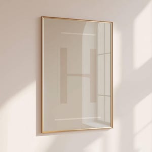 May include: A framed print with a gold-coloured frame. The print features a large, light brown letter "H" centered on a light beige background. Two horizontal lines are above and below the letter. The frame is rectangular and hangs on a light beige wall.