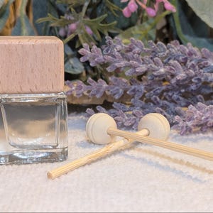 May include: A clear glass bottle with a light brown wooden cap, accompanied by two wooden diffuser sticks with round tops. The background features lavender sprigs and greenery, suggesting a fragrance product.