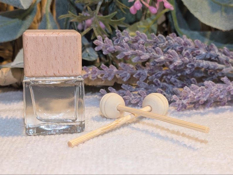 May include: A clear glass bottle with a light brown wooden cap, accompanied by two wooden diffuser sticks with round tops. The background features lavender sprigs and greenery, suggesting a fragrance product.