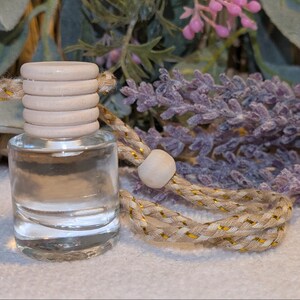 May include: A small, clear glass bottle with a wooden cap and a braided cord. The bottle is filled with a clear liquid. The cord is beige with gold accents. Lavender sprigs are in the background.