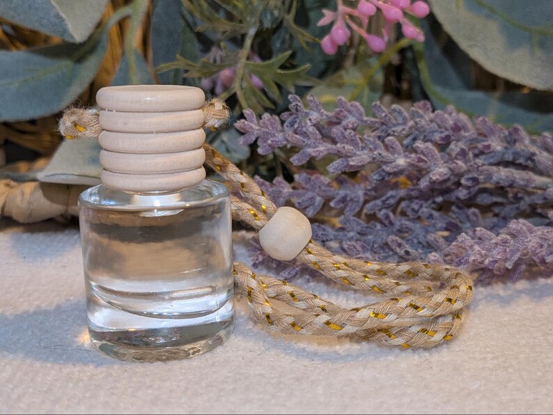 May include: A small, clear glass bottle with a wooden cap and a braided cord. The bottle is filled with a clear liquid. The cord is beige with gold accents. Lavender sprigs are in the background.