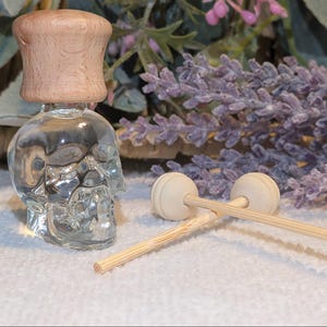 May include: A clear glass skull-shaped bottle with a light brown wooden cap. Two small wooden diffuser sticks and a lavender sprig are also visible. The bottle is likely for essential oils or fragrance.