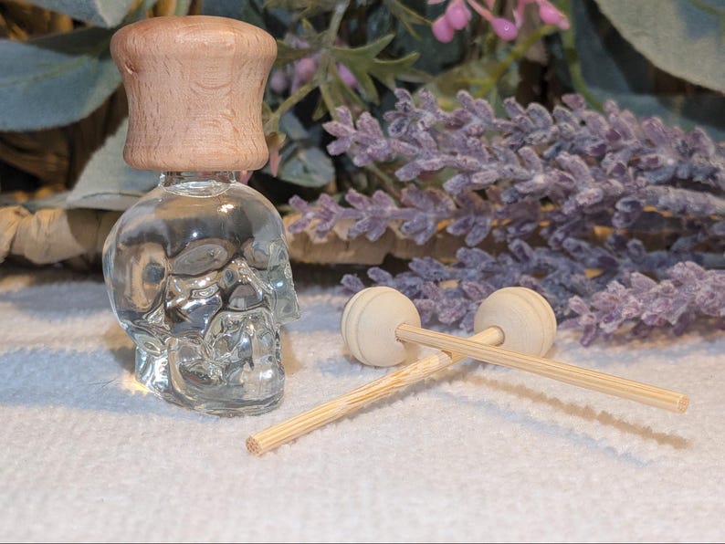 May include: A clear glass skull-shaped bottle with a light brown wooden cap. Two small wooden diffuser sticks and a lavender sprig are also visible. The bottle is likely for essential oils or fragrance.