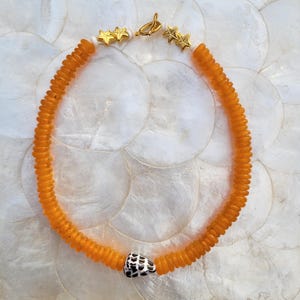 Peut inclure: Collier de perles orange avec des détails dorés en forme d'étoiles et un fermoir doré. Le collier comprend une perle à motifs noirs et blancs. Un accessoire élégant.