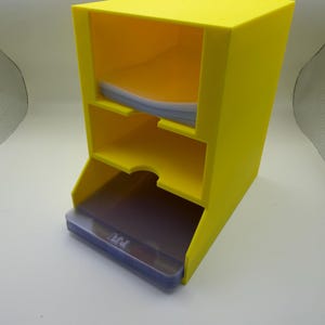 May include: A bright yellow, three-tiered storage organizer. The organizer has a slanted design with open compartments for storing items. The bottom compartment holds a stack of clear, plastic cards. The top two compartments are empty.