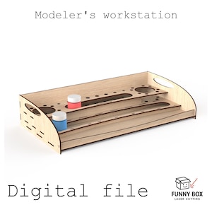Può includere: Un organizer in legno chiaro per postazione da modellismo con ripiani a gradini, fori circolari per barattoli di vernice e fessure per attrezzi. Il testo riporta Modelers workstation e Digital file con logo Funny Box.