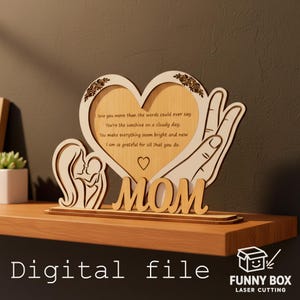 Pode incluir: Peça decorativa em madeira cortada a laser com moldura em forma de coração, silhueta de mãe e filho e a palavra "MOM". A moldura contém uma mensagem de amor. A base diz "Digital file" e o logotipo Funny Box Laser Cutting.