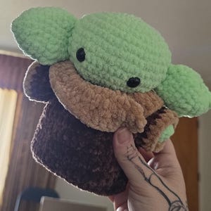 Puede incluir: Un peluche de ganchillo que representa a un personaje popular. El juguete tiene una cabeza y orejas de color verde claro, ojos negros y un atuendo marr&oacute;n y beige. El peluche est&aacute; hecho de un hilo suave y texturizado.