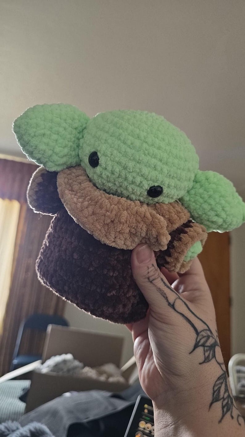 Puede incluir: Un peluche de ganchillo que representa a un personaje popular. El juguete tiene una cabeza y orejas de color verde claro, ojos negros y un atuendo marr&oacute;n y beige. El peluche est&aacute; hecho de un hilo suave y texturizado.