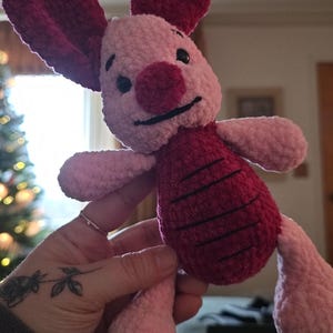 Puede incluir: Un peluche hecho a mano de un personaje con colores rosa y burdeos. El juguete tiene grandes orejas burdeos, una nariz burdeos y una barriga burdeos con rayas negras. El juguete est&aacute; siendo sostenido por una persona.