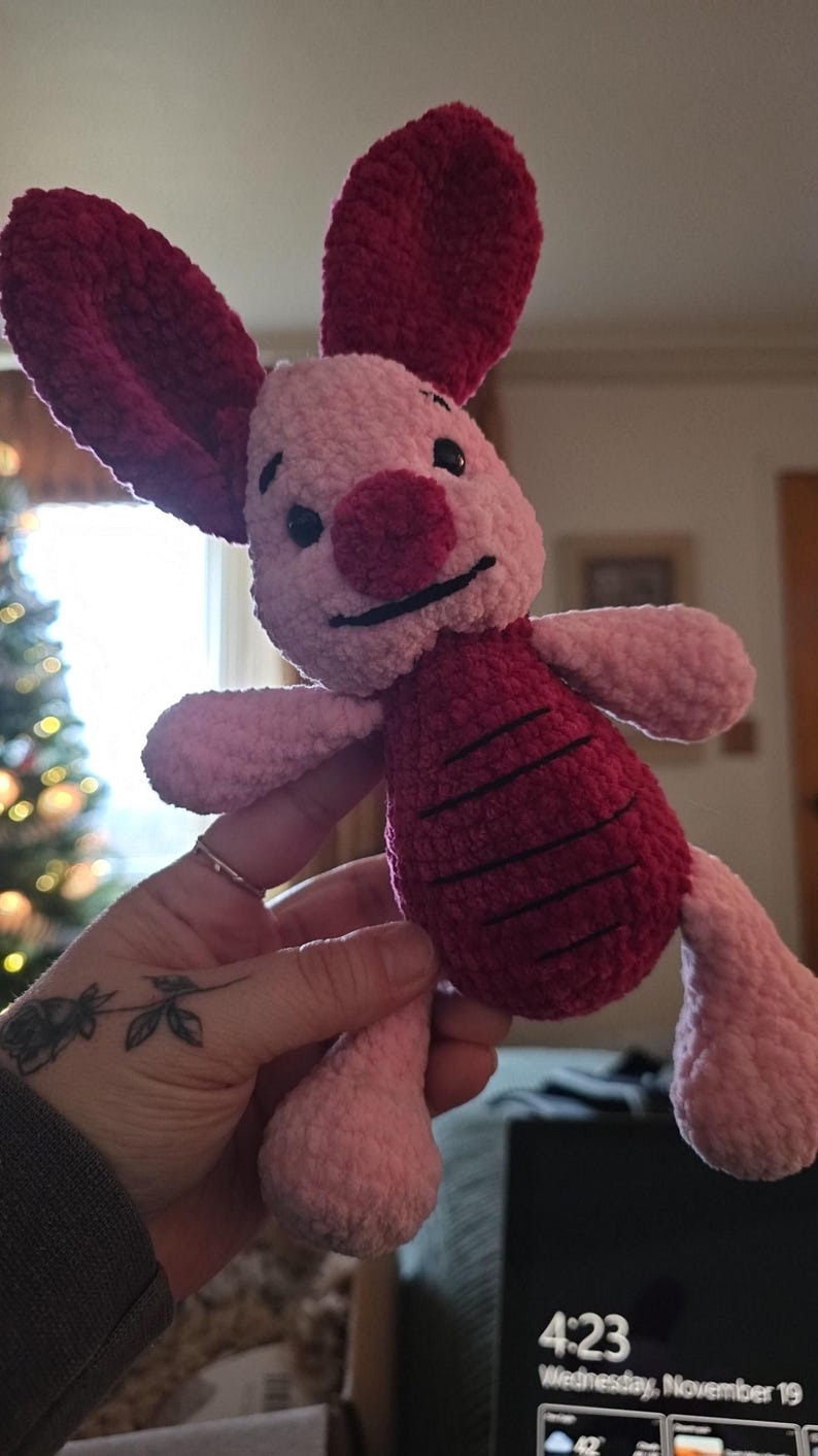 Puede incluir: Un peluche hecho a mano de un personaje con colores rosa y burdeos. El juguete tiene grandes orejas burdeos, una nariz burdeos y una barriga burdeos con rayas negras. El juguete est&aacute; siendo sostenido por una persona.