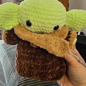 Puede incluir: Un peluche hecho a mano de un personaje con orejas grandes y ojos negros grandes. El juguete es principalmente verde, marr&oacute;n y beige, con un aspecto suave y texturizado. El juguete est&aacute; sostenido en la mano de una persona.