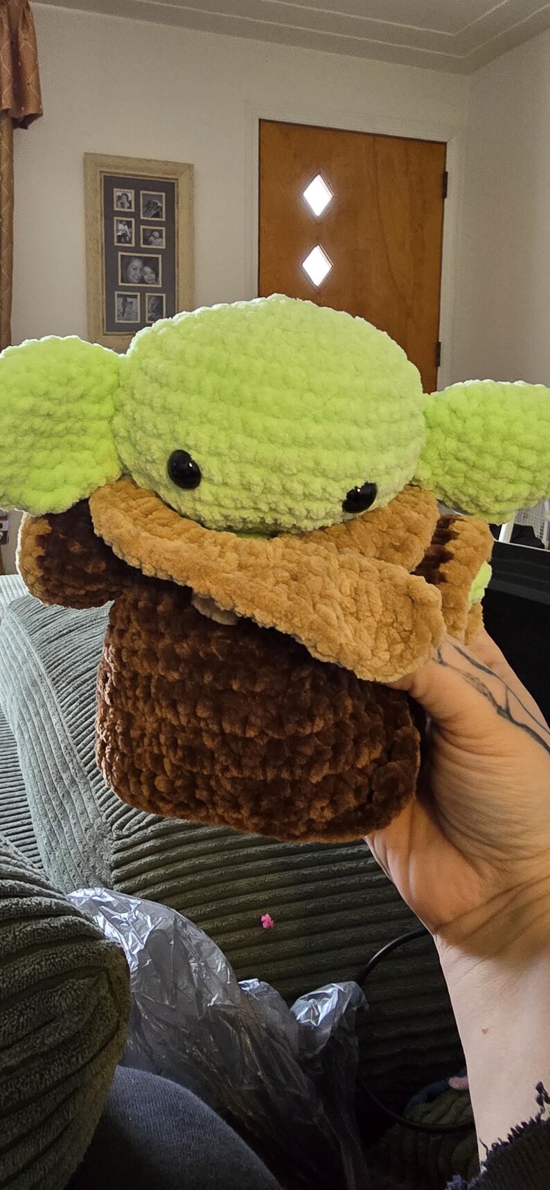 Puede incluir: Un peluche hecho a mano de un personaje con orejas grandes y ojos negros grandes. El juguete es principalmente verde, marr&oacute;n y beige, con un aspecto suave y texturizado. El juguete est&aacute; sostenido en la mano de una persona.