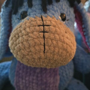 Puede incluir: Peluche de Eeyore hecho a mano con ganchillo. El burro tiene un cuerpo azul claro, un hocico marr&oacute;n y pelo negro. El juguete presenta una barriga morada y orejas internas rosas. Personaje infantil.