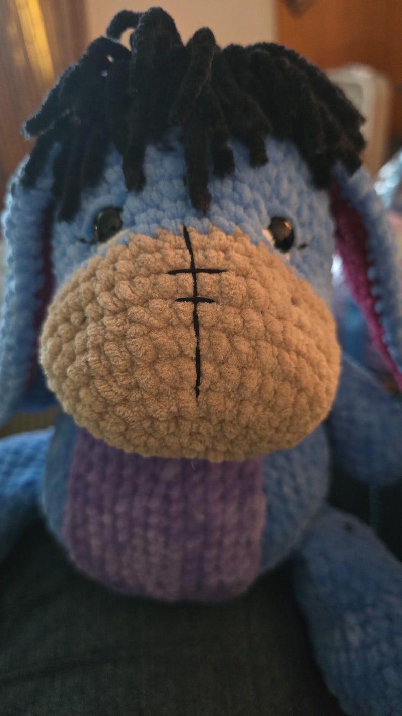 Puede incluir: Peluche de Eeyore hecho a mano con ganchillo. El burro tiene un cuerpo azul claro, un hocico marr&oacute;n y pelo negro. El juguete presenta una barriga morada y orejas internas rosas. Personaje infantil.