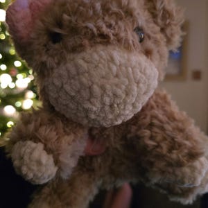 Puede incluir: Un peluche de vaca con cuerpo marr&oacute;n claro, hocico blanco y lazo rosa. El texto "Fluffy cow.... she came out so cute!" est&aacute; en la parte inferior de la imagen. El juguete tiene un aspecto suave y texturizado.