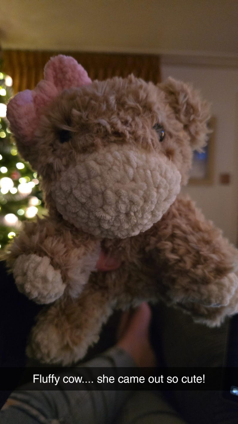 Puede incluir: Un peluche de vaca con cuerpo marr&oacute;n claro, hocico blanco y lazo rosa. El texto "Fluffy cow.... she came out so cute!" est&aacute; en la parte inferior de la imagen. El juguete tiene un aspecto suave y texturizado.