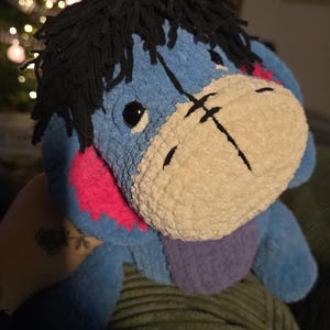 Puede incluir: Un peluche de Eeyore, el burro de Winnie the Pooh, en tonos azules, rosas y morados. El juguete tiene pelo de hilo negro y un hocico color crema. El juguete es suave y tierno.