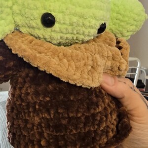 Puede incluir: Mu&ntilde;eco de ganchillo hecho a mano, que se asemeja a un personaje de una franquicia de ciencia ficci&oacute;n. El mu&ntilde;eco tiene una cabeza y orejas de color verde claro, un cuello y pecho de color beige y un cuerpo marr&oacute;n oscuro. El mu&ntilde;eco tiene grandes ojos negros.