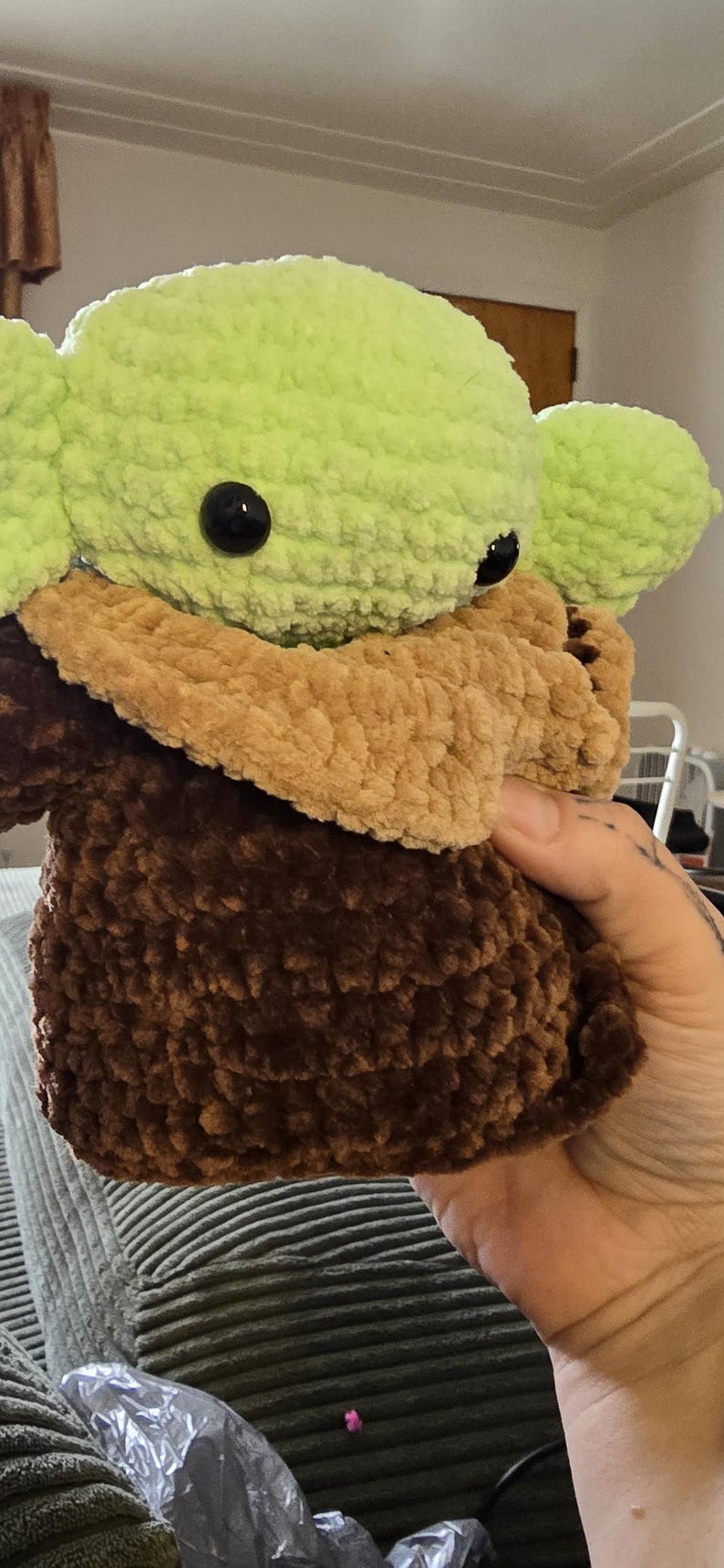Puede incluir: Mu&ntilde;eco de ganchillo hecho a mano, que se asemeja a un personaje de una franquicia de ciencia ficci&oacute;n. El mu&ntilde;eco tiene una cabeza y orejas de color verde claro, un cuello y pecho de color beige y un cuerpo marr&oacute;n oscuro. El mu&ntilde;eco tiene grandes ojos negros.