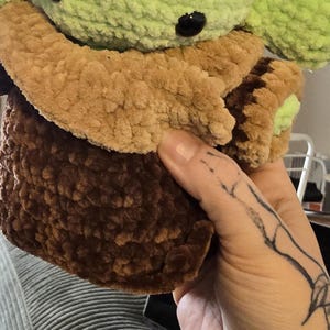 Puede incluir: Un peluche hecho a mano de un personaje con grandes ojos negros y orejas grandes. El juguete tiene una cabeza y orejas de color verde claro, un abrigo de color canela y un cuerpo marr&oacute;n oscuro. El juguete est&aacute; hecho de un material suave y texturizado.