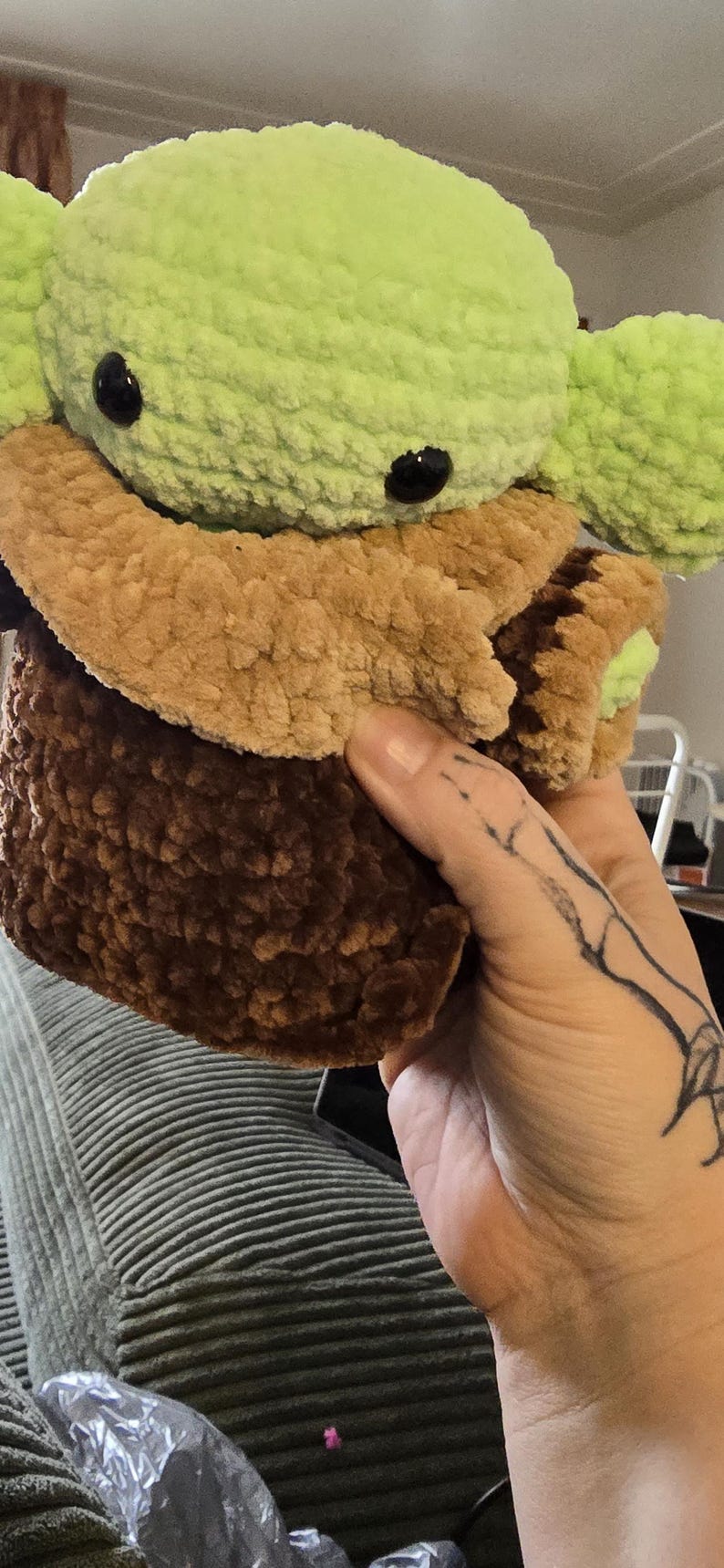 Puede incluir: Un peluche hecho a mano de un personaje con grandes ojos negros y orejas grandes. El juguete tiene una cabeza y orejas de color verde claro, un abrigo de color canela y un cuerpo marr&oacute;n oscuro. El juguete est&aacute; hecho de un material suave y texturizado.