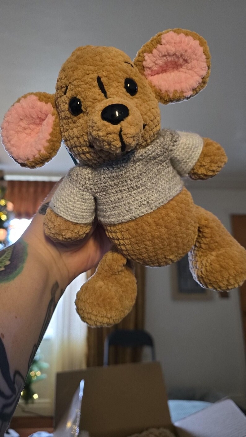 Puede incluir: Un juguete de peluche hecho a mano con forma de oso. El oso es marr&oacute;n con el interior de las orejas rosa y un su&eacute;ter gris. Tiene ojos negros y una nariz negra. El juguete es suave y tierno.