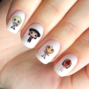 May include: Four white nail decals with different characters from popular horror movies. The decals feature Beetlejuice, Wednesday Addams, the Headless Horseman, and the Cenobite from Hellraiser.