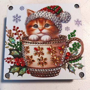 May include: A square, white Christmas-themed decoration featuring a ginger kitten wearing a Santa hat, nestled in a decorated teacup. The teacup and hat are embellished with rhinestones. Holly and snowflake accents complete the festive design.